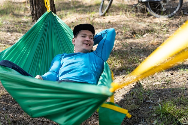 Junger glücklicher lächelnder Mann im Hut, der draußen in der Hängematte im Wald entspannt