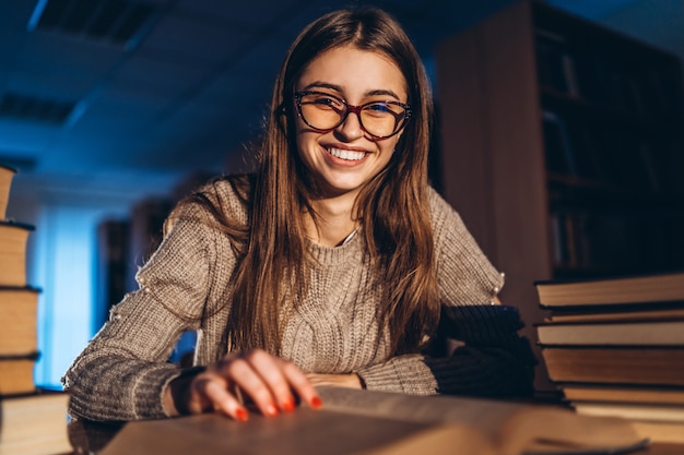 Junger glücklicher lächelnder Student in den Gläsern, die sich auf die Prüfung vorbereiten. Mädchen am Abend sitzt an einem Tisch in der Bibliothek mit einem Stapel Bücher