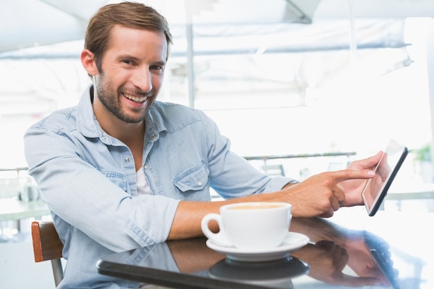 Junger glücklicher Mann, der an der Kamera lächelt und auf das Telefon im Café zeigt