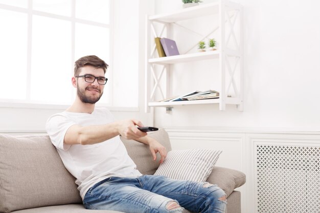 Junger glücklicher Mann, der auf der Couch fernsieht und mit der Fernbedienung auf den Fernseher zeigt, Platz kopieren