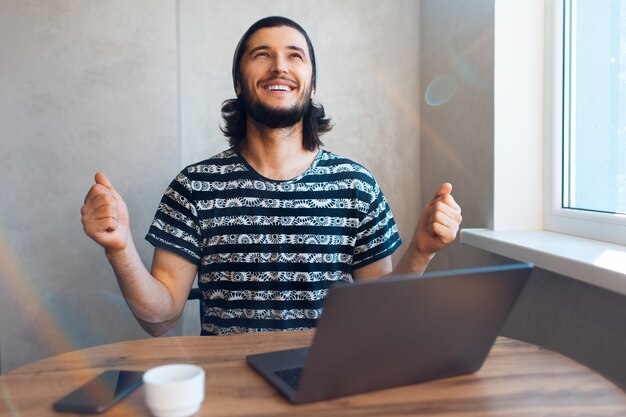 Junger glücklicher Mann, der sehr glücklich an seinem Laptop mit Kaffee und Smartphone auf dem Tisch arbeitet