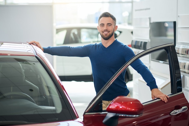 Junger glücklicher Mann, der sich auf Auto im Autohaus stützt.