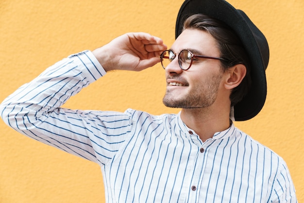 junger glücklicher Mann mit Brille und schwarzem Hut, der lächelt und beiseite auf den Kopienraum schaut, der über gelber Wand isoliert ist?