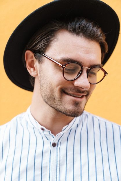 Foto junger glücklicher mann mit brille und schwarzem hut lächelt und schaut nach oben auf den kopienraum, der über gelber wand isoliert ist?