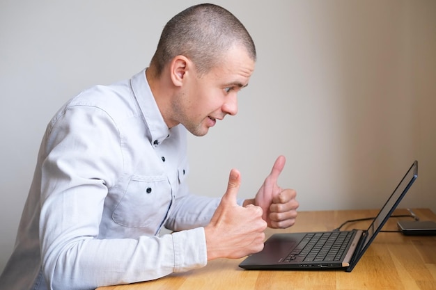 Junger glücklicher Mann zeigt Geste wie oder Daumen nach oben, während er am Laptop arbeitet oder einkauft