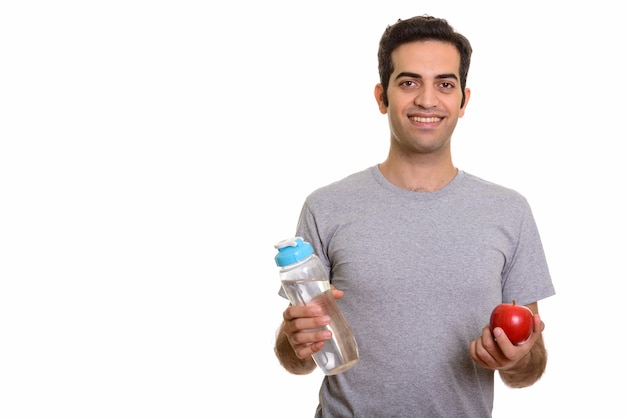 Junger glücklicher persischer Mann, der Wasserflasche und roten Apfel bereit für Turnhalle hält