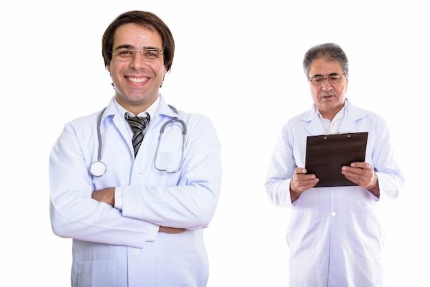 Junger glücklicher persischer Mannarzt, der mit verschränkten Armen lächelt, und älterer Arzt, der Zwischenablage betrachtet