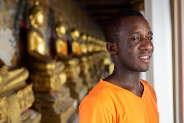 Junger glücklicher Touristenmann, der lächelt, während er gegen Ansicht der ausgerichteten Buddha-Strukturen im Wat Arun-Tempel bei Bangkok Thailand denkt