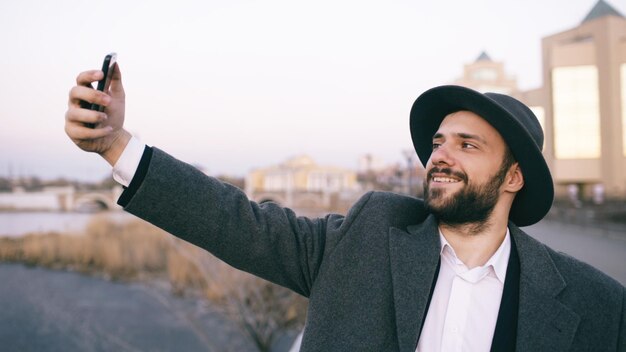 Junger glücklicher touristischer Mann lächelt, während er Selfie-Foto mit Handy am Flussufer der Stadt macht