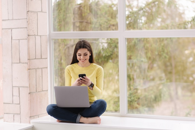 Junger Grafikdesigner, der zu Hause arbeitet. Frau, die auf Fensterbank mit Laptop sitzt und Kaffee trinkt.