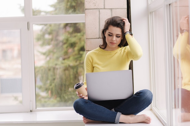 Junger Grafikdesigner, der zu Hause arbeitet. Frau, die auf Fensterbank mit Laptop sitzt und Kaffee trinkt.