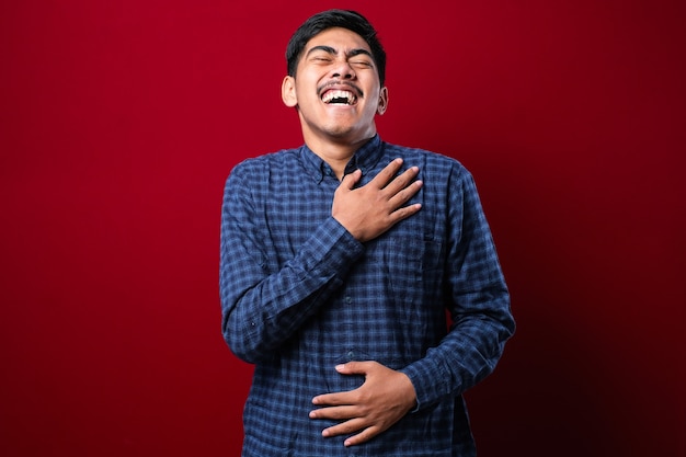 Junger gutaussehender asiatischer Junge, der ein lässiges Hemd trägt, das über isoliertem rotem Hintergrund steht und lächelt und laut lacht, weil lustiger verrückter Witz mit den Händen am Körper.