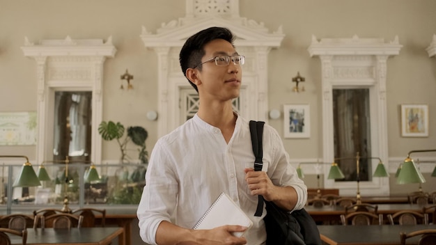 Junger gutaussehender asiatischer männlicher Student mit Rucksack, der selbstbewusst in die Universitätsbibliothek kommt Attraktiver Kerl mit Brille, der am College studiert