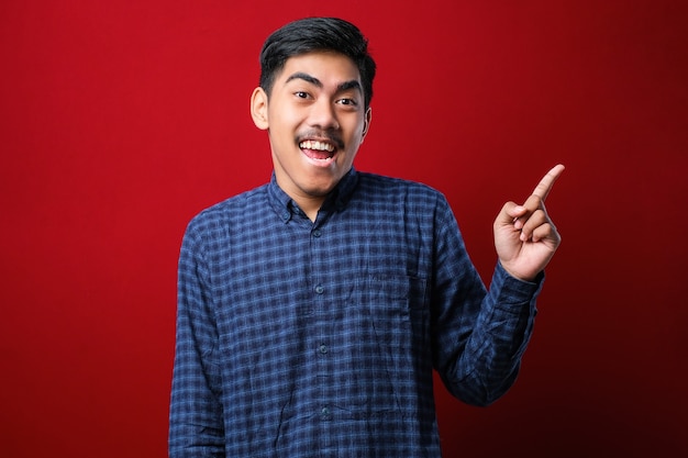Junger gutaussehender asiatischer Mann, der ein lässiges Hemd über rotem Hintergrund mit einem großen Lächeln im Gesicht trägt und mit dem Handfinger auf die Seite zeigt, die in die Kamera schaut.