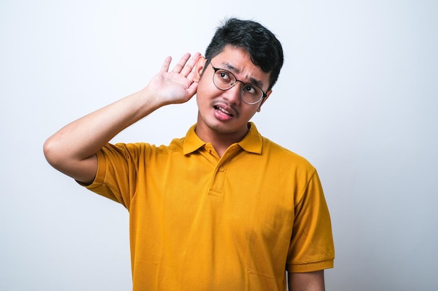 Junger gutaussehender asiatischer Mann mit legerem Hemd, der vor isoliertem weißem Hintergrund steht und mit der Hand über dem Ohr lächelt und einer Anhörung zu Gerüchten oder Klatsch zuhört Taubheitskonzept