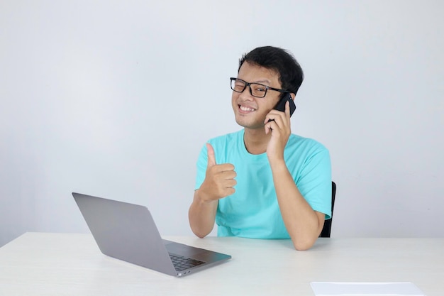 Junger gutaussehender asiatischer Mann zeigt Daumen nach oben, während er telefoniert und mit Laptop arbeitet Indonesischer Mann mit blauem Hemd