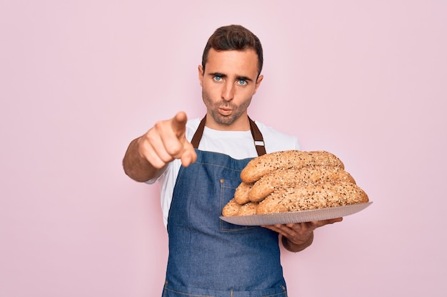 Junger, gutaussehender Bäckermann mit blauen Augen, der eine Schürze trägt und ein Tablett mit Vollkornbrot hält, mit dem Finger in die Kamera zeigt und Ihnen von vorne ein positives und selbstbewusstes Handzeichen zeigt