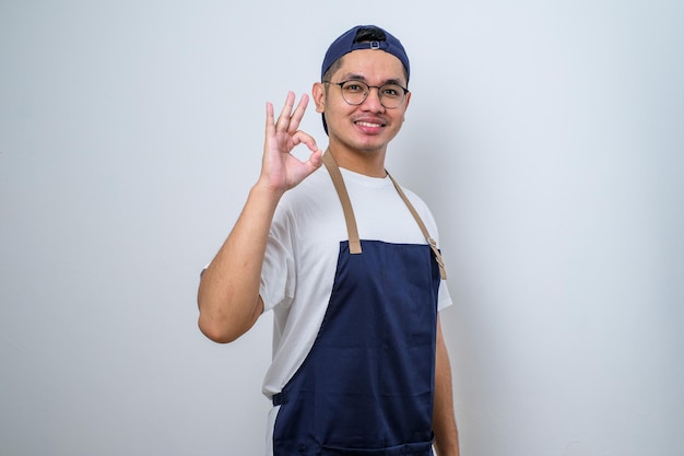 Junger gutaussehender Barista-Mann mit Schürze lächelt und macht mit den Fingern ein ausgezeichnetes Symbol