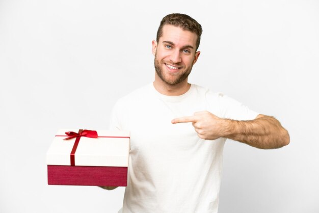 Junger gutaussehender blonder Mann, der ein Geschenk über isoliertem weißem Hintergrund hält und darauf zeigt
