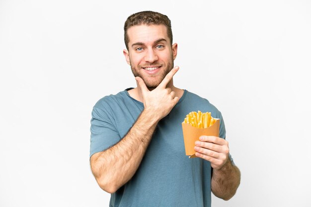 Junger gutaussehender blonder Mann, der gebratene Chips über isoliertem weißem Hintergrund hält, glücklich und lächelnd