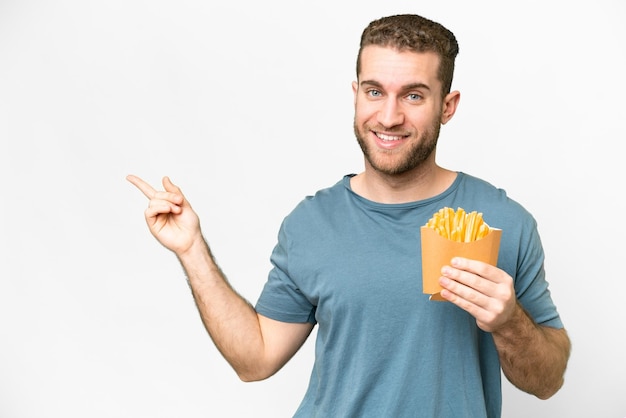Junger gutaussehender blonder Mann, der gebratene Chips über isoliertem weißem Hintergrund hält und mit dem Finger zur Seite zeigt