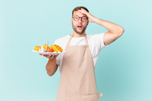 Junger gutaussehender blonder Mann, der glücklich, erstaunt und überrascht aussieht. Waffeln kochen Konzept