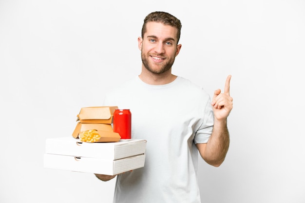 Junger gutaussehender blonder Mann, der Pizzen und Burger vor isoliertem Hintergrund hält und einen Finger im Zeichen der Besten zeigt und hebt