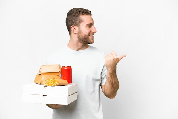 Junger gutaussehender blonder Mann, der Pizzen und Burger vor isoliertem Hintergrund hält und zur Seite zeigt, um ein Produkt zu präsentieren