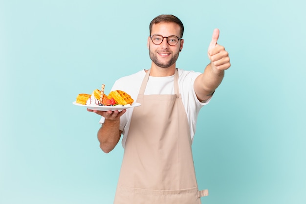 Junger gutaussehender blonder Mann, der stolz ist und positiv mit Daumen nach oben lächelt. Waffeln kochen Konzept