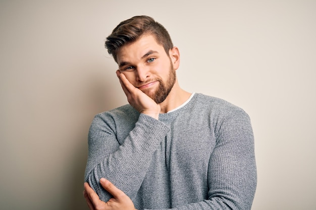 Junger gutaussehender blonder Mann mit Bart und blauen Augen trägt einen lässigen Pullover und sieht müde und gelangweilt aus, mit Depressionsproblemen und verschränkten Armen