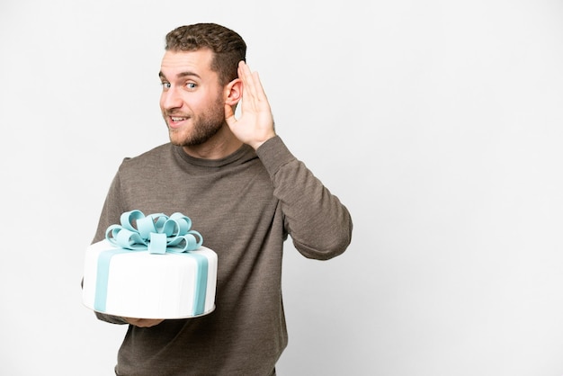 Junger gutaussehender blonder Mann mit einem großen Kuchen vor isoliertem weißem Hintergrund, der etwas hört, indem er die Hand auf das Ohr legt