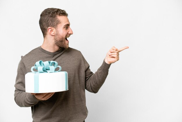 Junger gutaussehender blonder Mann mit einem großen Kuchen vor isoliertem weißem Hintergrund, der mit dem Finger zur Seite zeigt und ein Produkt präsentiert