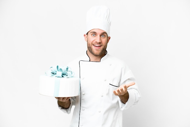 Junger gutaussehender blonder Mann mit einem großen Kuchen vor isoliertem weißem Hintergrund mit schockiertem Gesichtsausdruck