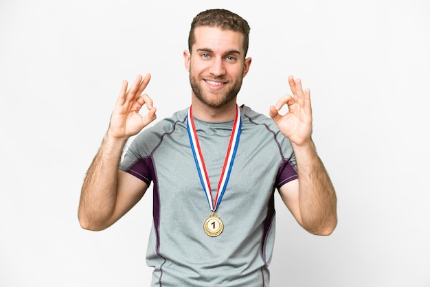 Foto junger gutaussehender blonder mann mit medaillen vor isoliertem weißem hintergrund, der mit den fingern ein ok-zeichen zeigt