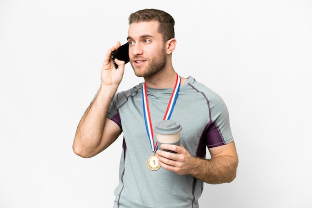 Junger gutaussehender blonder Mann mit Medaillen vor isoliertem weißem Hintergrund mit Kaffee zum Mitnehmen und einem Handy