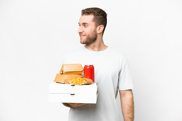 Junger gutaussehender blonder Mann mit Pizzen und Burgern vor isoliertem Hintergrund mit Blick auf die Seite