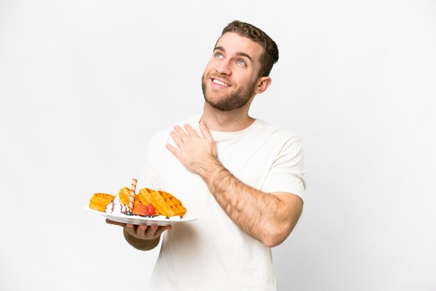 Junger gutaussehender blonder Mann mit Waffeln vor isoliertem weißem Hintergrund, der lächelnd aufblickt