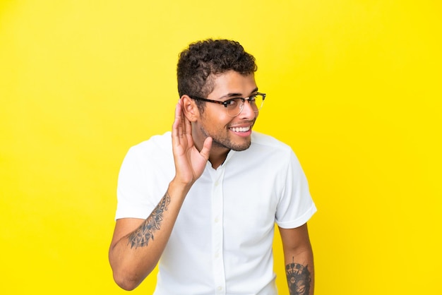 Junger gutaussehender Brasilianer isoliert auf gelbem Hintergrund, der etwas hört, indem er die Hand auf das Ohr legt
