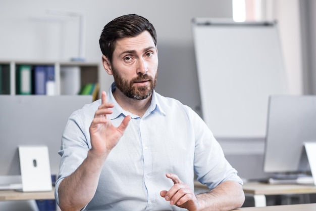 Junger gutaussehender erfolgreicher Business Coach erklärt und erzählt Kameragesten mit den Händen Sitzt an einem Computertisch im Büro und schaut in die Kamera