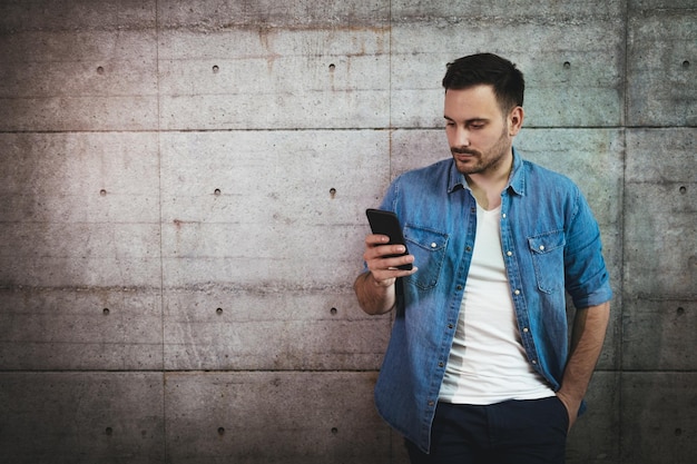 Junger gutaussehender erfolgreicher Unternehmer, der eine Nachricht auf einem Smartphone vor einer Betonwand schreibt.