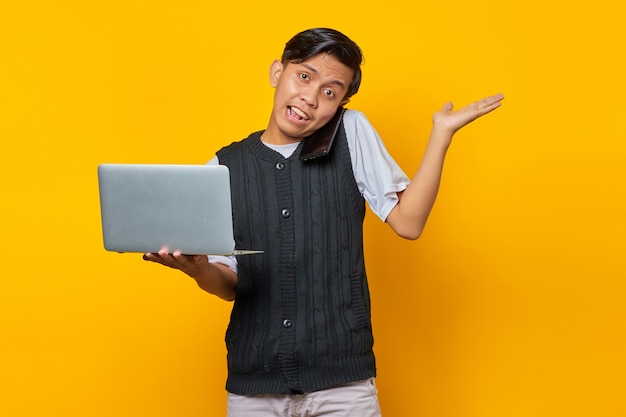 Junger gutaussehender Geschäftsmann, der auf einem Smartphone spricht, während er einen Laptop auf gelbem Hintergrund hält