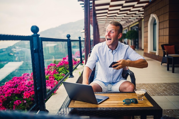 Junger gutaussehender Geschäftsmann in einem weißen Hemd und kurzen Hosen sitzt mit einem Laptop in einem Café am Tisch Arbeiten, während er sich ausruht