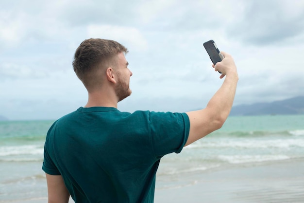 Junger gutaussehender glücklicher Mann auf See fotografiert sich selbst mit der Kamera seines Telefons mit Smartphone für soziale Medien am Sommerstrand in tropischem exotischem Land Blogger Vlog
