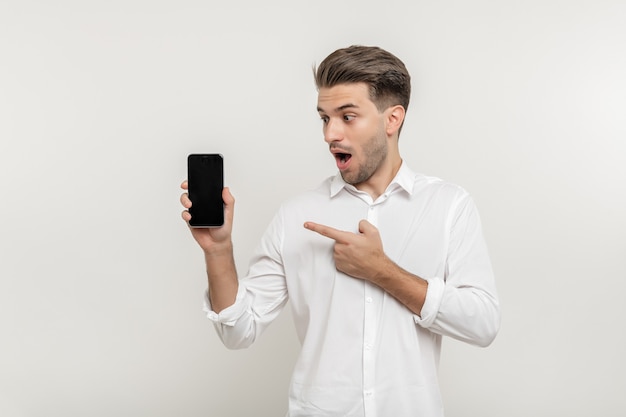 Foto junger gutaussehender glücklicher mann, der ein smartphone mit leerem bildschirm hält und isoliert auf das handy zeigt und beobachtet