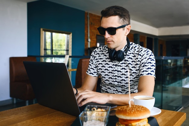 Junger gutaussehender Hipster-Mann mit Sonnenbrille benutzt Laptop in der Cafeteria-Tasse Kaffee und Cheeseburger