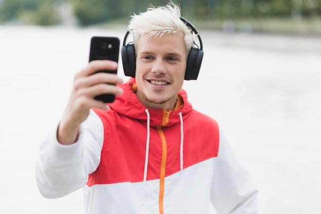 Junger gutaussehender Hipster-Mann mit trendigem Reißverschluss-Hoodie und Musikhören mit Bluetooth-Kopfhörern hat Spaß beim Selbstporträt mit Smartphone im Freien Lifestyle-Technologiekonzept für Menschen
