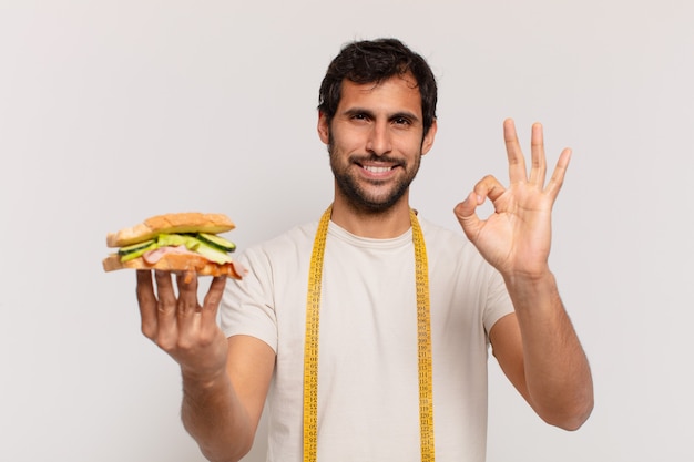 Junger gutaussehender indischer Mann glücklicher Ausdruck und hält ein Sandwich