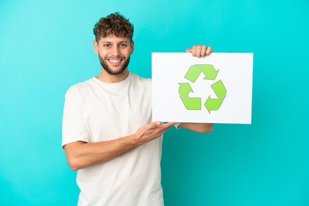 Junger gutaussehender kaukasischer Mann isoliert auf blauem Hintergrund, der ein Plakat mit Recycling-Symbol mit glücklichem Ausdruck hält