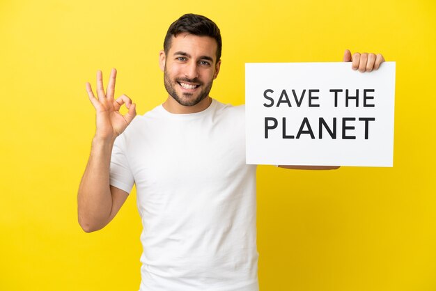 Junger gutaussehender kaukasischer Mann isoliert auf gelbem Hintergrund, der ein Plakat mit dem Text Save the Planet hält und einen Sieg feiert