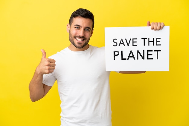 Junger gutaussehender kaukasischer Mann isoliert auf gelbem Hintergrund, der ein Plakat mit dem Text Save the Planet mit Daumen nach oben hält